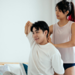Woman assists man in a shoulder stretch at home, with a bed and bright window in the background.