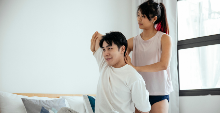 Woman assists man in a shoulder stretch at home, with a bed and bright window in the background.