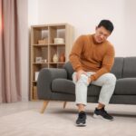 Man seated on a gray sofa in a living room, wincing and touching his knee.