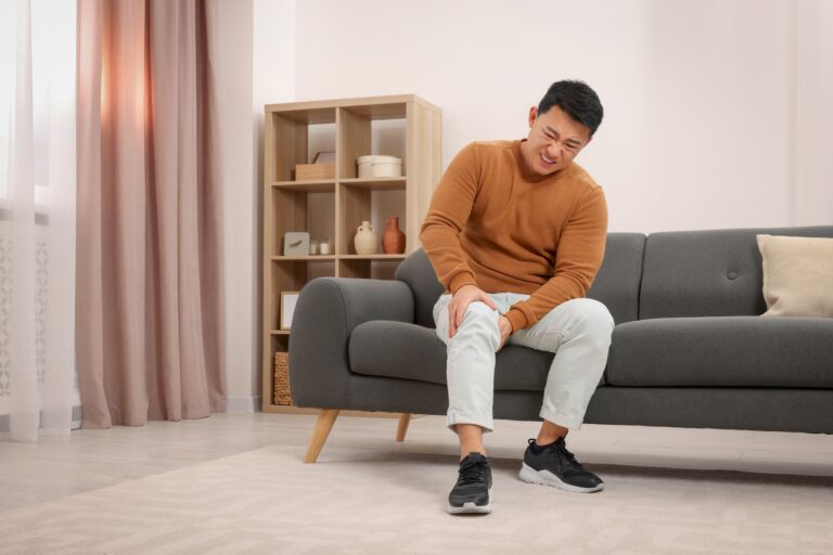 Man seated on a gray sofa in a living room, wincing and touching his knee.